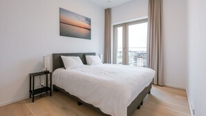 Ett modernt sovrum med en stor säng, ett sängbord, en liten avlastningsyta och utsikt över staden genom ett stort fönster.