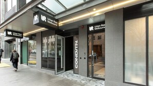 Een gebouw met een bord waarop 'Urban Rest Melbourne' staat, dat een hotel of accommodatie aanduidt. Er is een glazen deur en een zichtbaar parkeergedeelte.