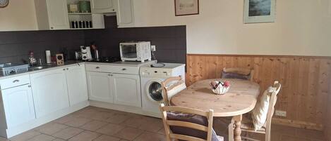 A compact kitchen with white cabinets, a microwave, and a washing machine. There is a wooden dining table with chairs and a small decorative bowl.