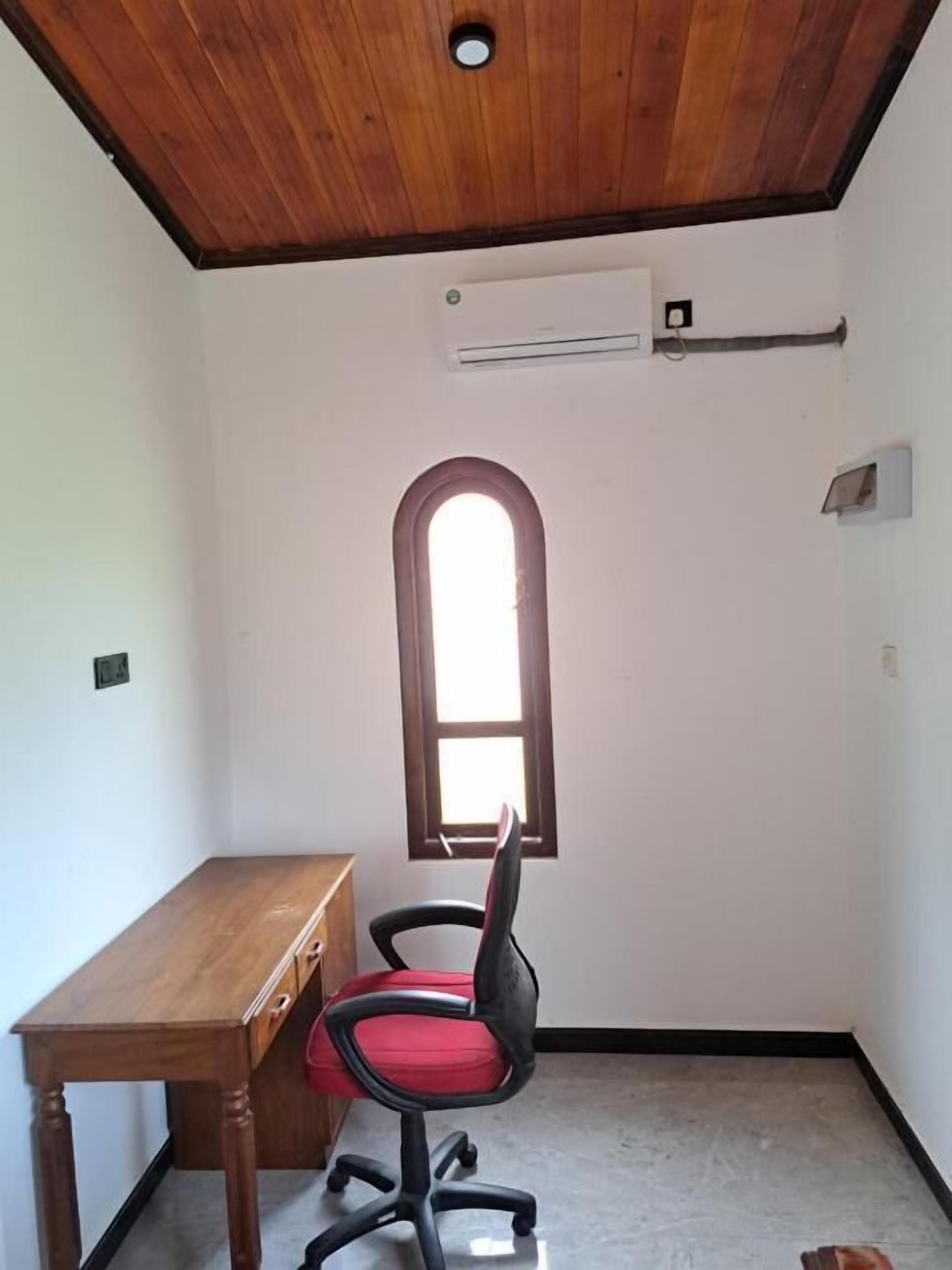 A room with a wooden ceiling, a desk, a red office chair, a window, and an air conditioning unit.