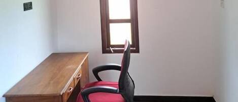 A room with a wooden ceiling, a desk, a red office chair, a window, and an air conditioning unit.