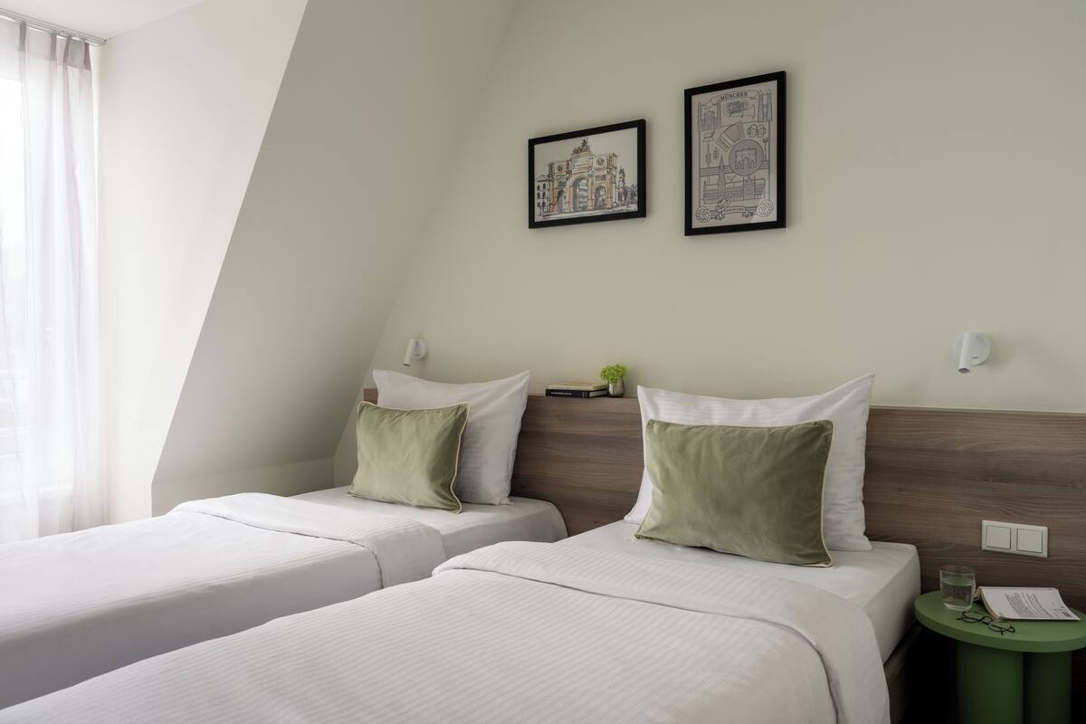 Une chambre d’hôtel avec deux lits, une petite table sur laquelle se trouve un verre d’eau, et deux tableaux encadrés accrochés au mur.