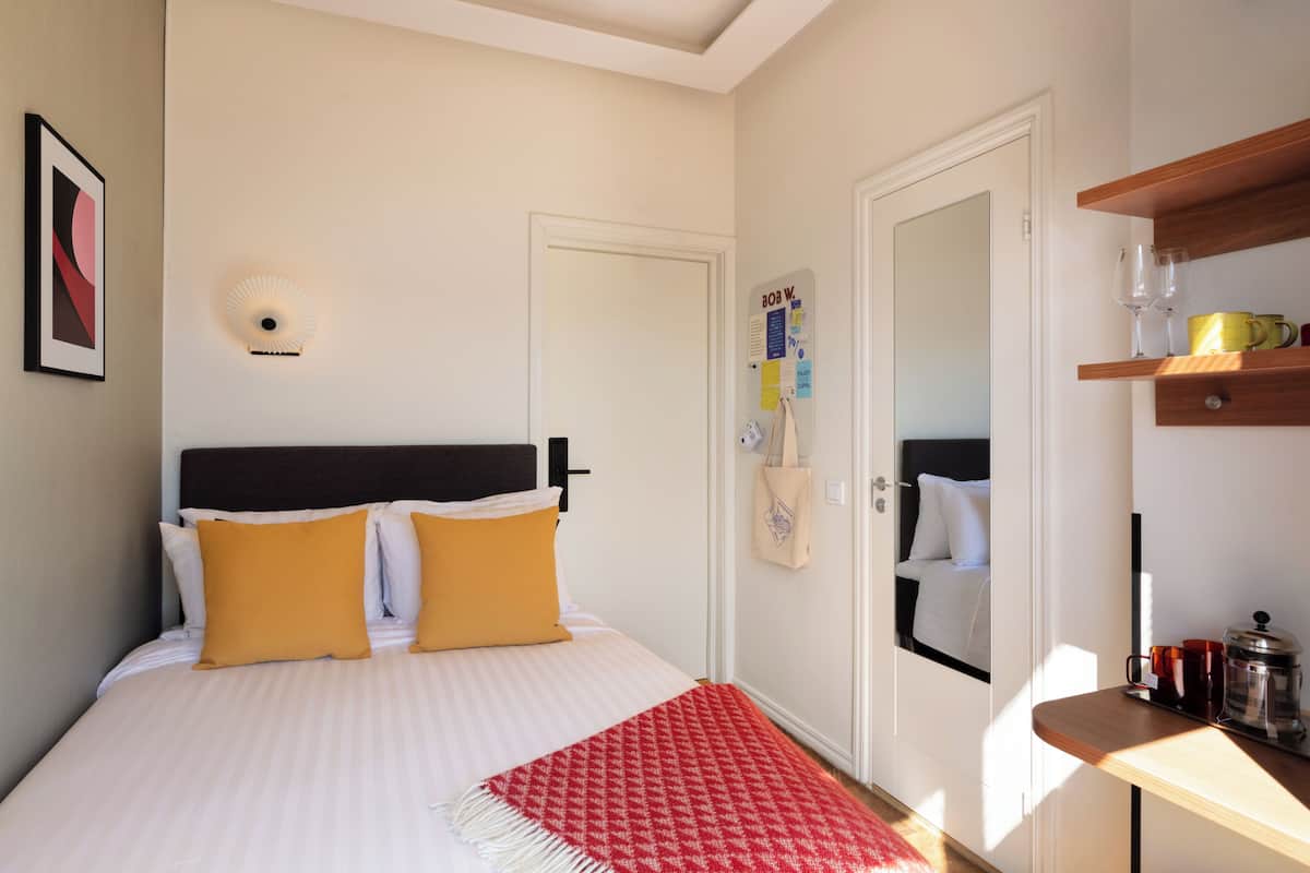 Une chambre d’hôtel comprenant un lit, une table de chevet avec une machine à café, un miroir et une porte sur laquelle est fixé un tableau d’affichage.