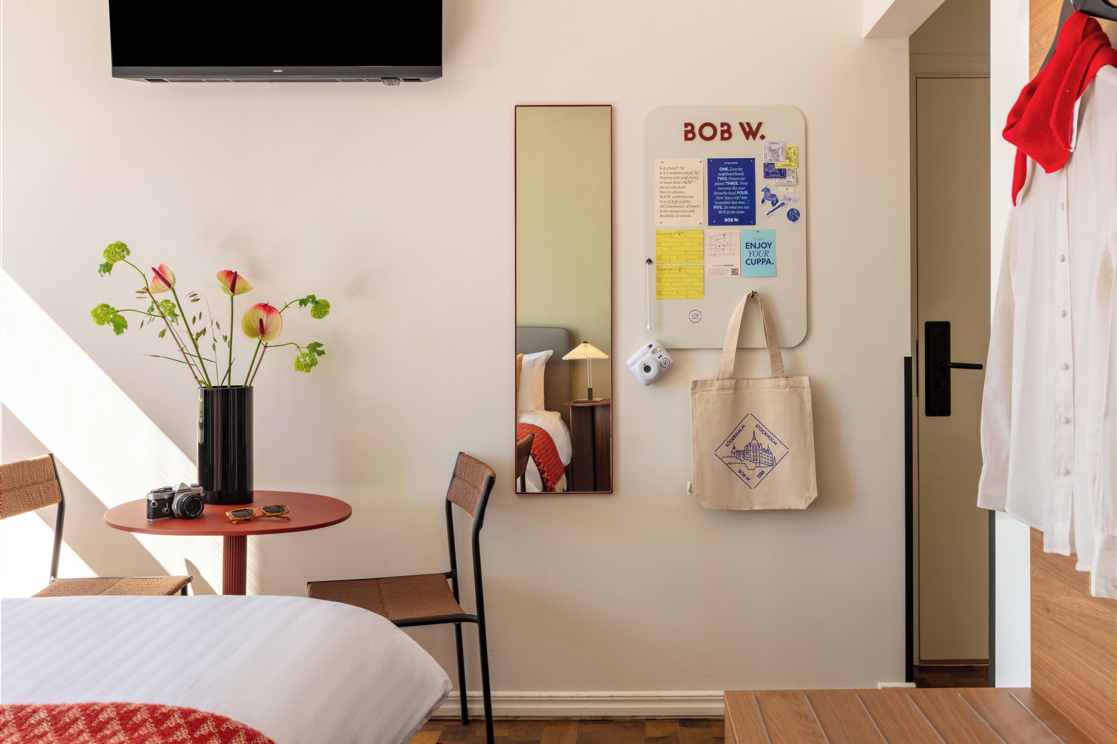 A hotel room with a bed, a small table with a camera and sunglasses, a chair, a mirror, a bulletin board with notes, and a TV mounted on the wall.