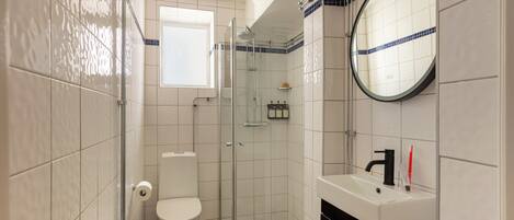 Un baño con inodoro, ducha con mampara de vidrio, lavamanos con espejo y ventana.