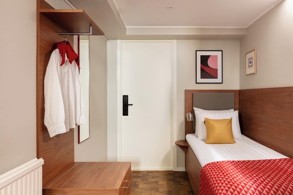 Une chambre d’hôtel avec un lit, une table de chevet en bois, une porte blanche et un porte-manteau sur lequel est accroché un vêtement rouge.