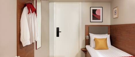 Habitación de hotel con cama, mesita de noche de madera, puerta blanca y perchero con una prenda roja.