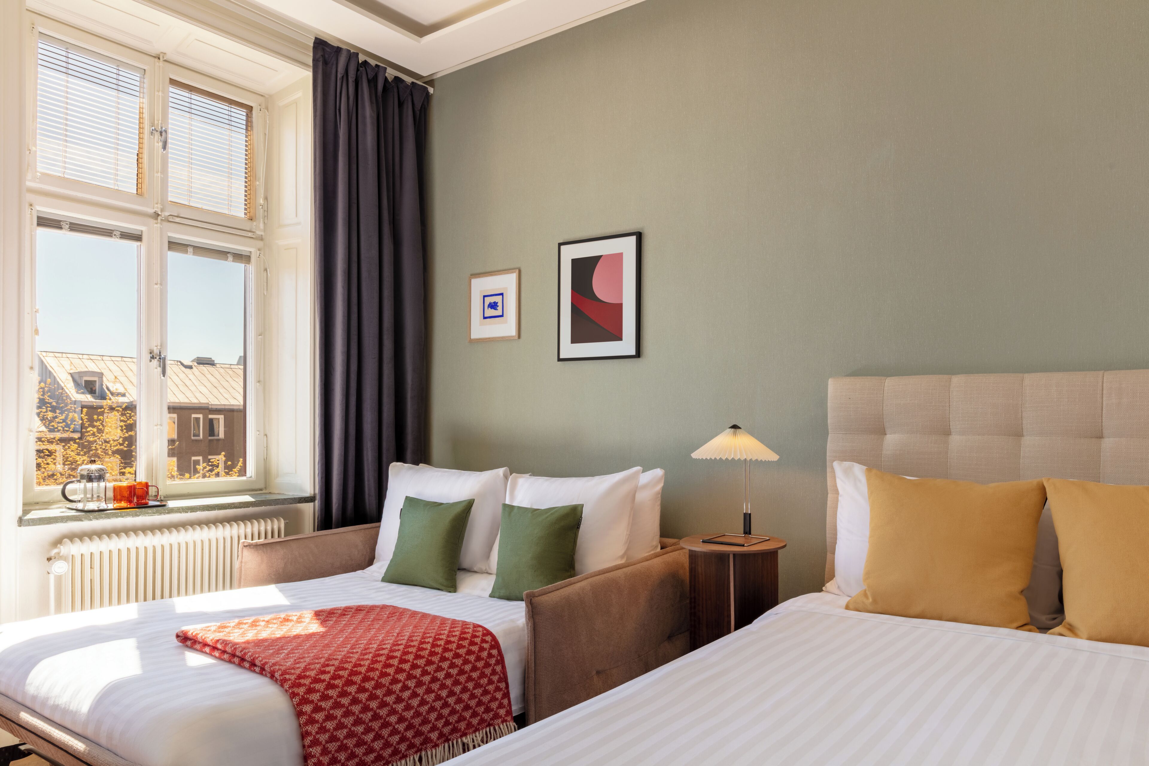 A hotel room with two beds, a window with blinds, a radiator, a lamp, and two framed pictures on the wall.