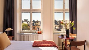 A hotel room with a bed, a table with a vase, a chair, and a window with a view of buildings. - Bob W Stockholm Sodermalm (Stockholm)