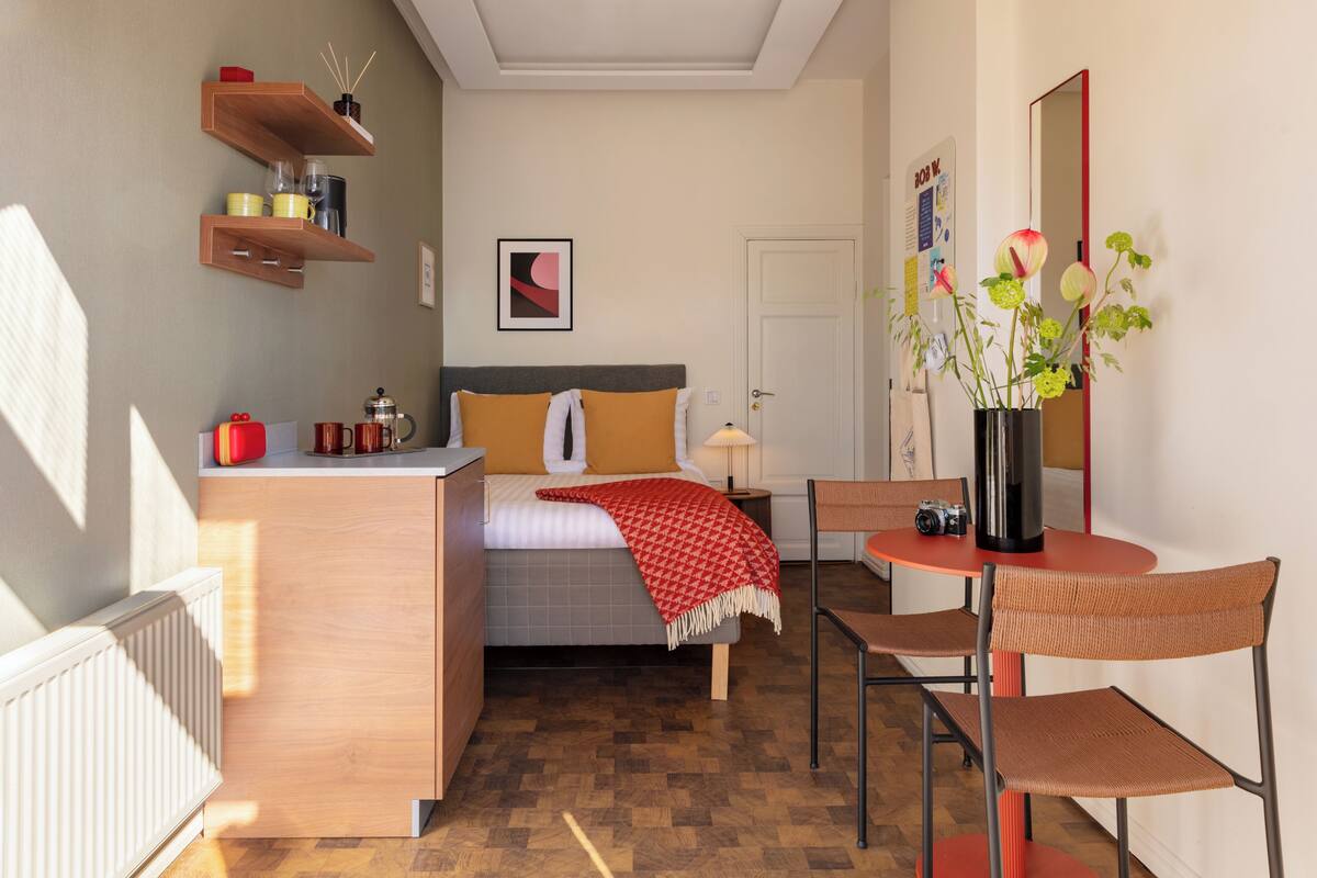 Une petite chambre comprenant un lit, une table de chevet, une chaise et une petite table sur laquelle est posé un vase de fleurs.