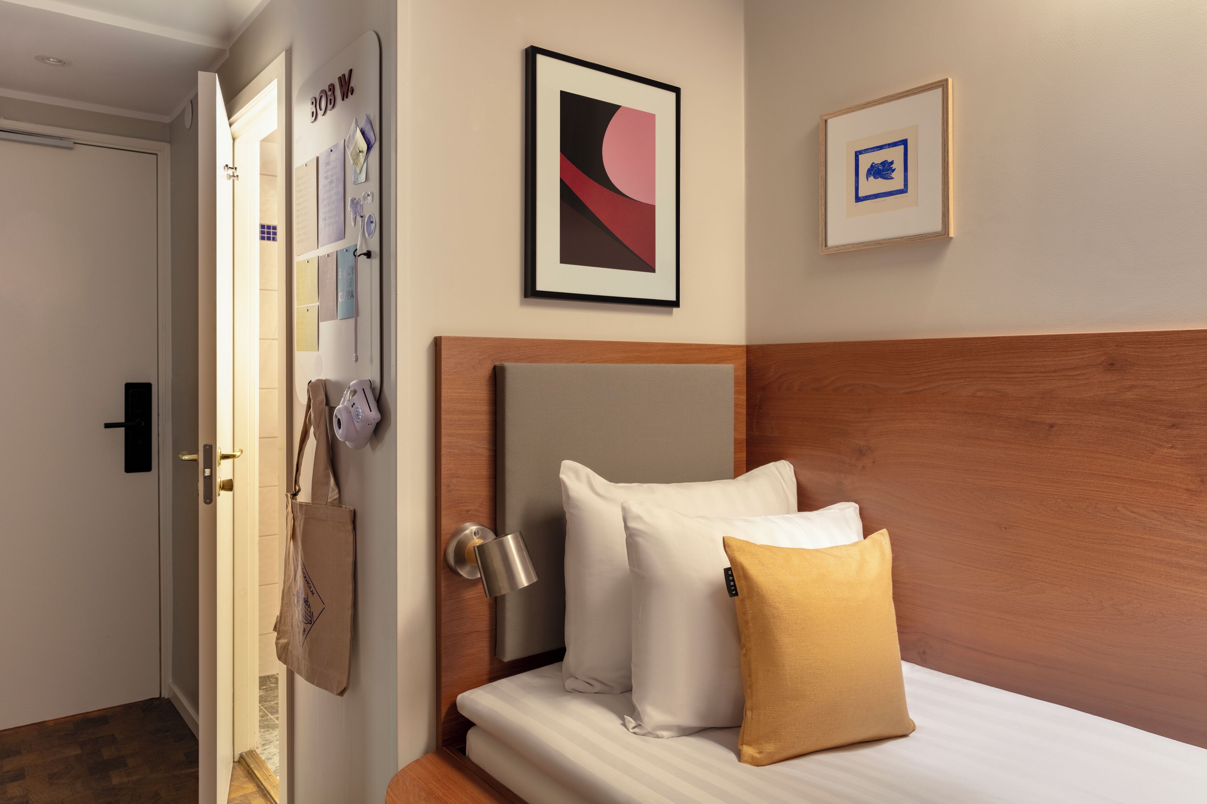 A hotel room with a bed, a wooden headboard, a wall-mounted lamp, a framed abstract painting, and a door with a bulletin board.