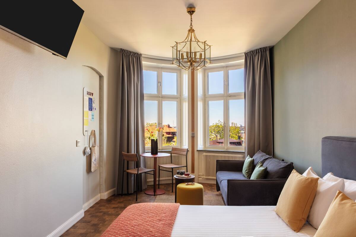Une chambre d’hôtel comprenant un lit, un canapé, une petite table avec des chaises, une fenêtre avec des rideaux et un lustre.