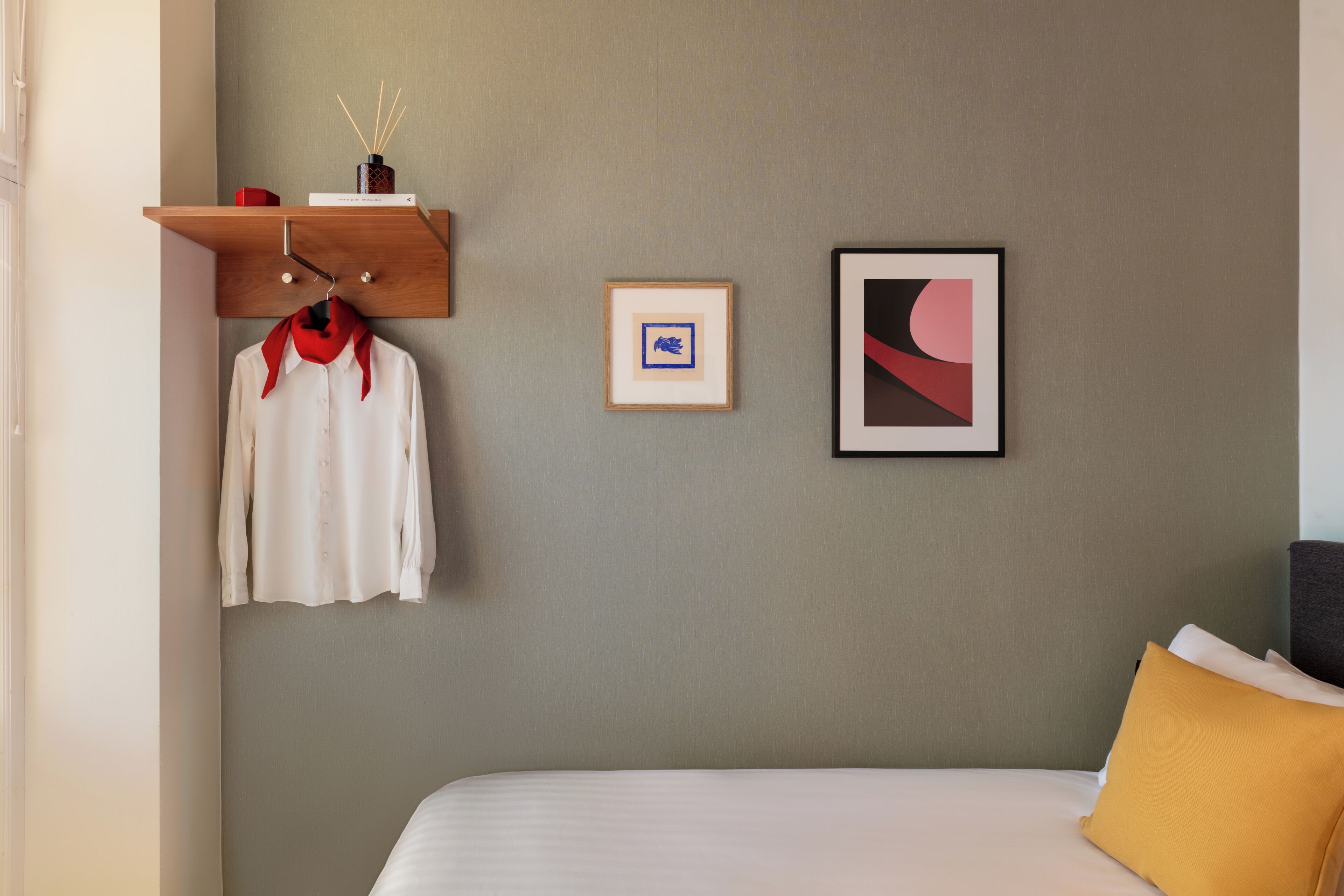 Ein Hotelzimmer mit einem Bett, einem roten Schal, einem gerahmten Bild und einem Regal mit Vase und Kerzen.