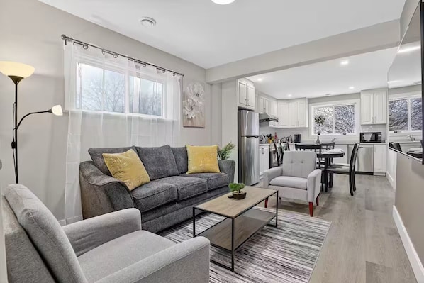 Ein modernes Wohnzimmer mit grauem Sofa, einem Couchtisch und einem Essbereich mit weißem Stuhl und einem kleinen Tisch.