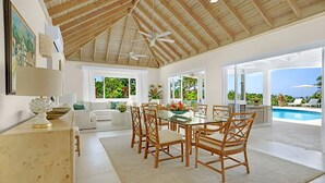 A spacious dining area with a glass table and wooden chairs, large windows offering views of greenery and a pool. - Tranquil 4BR Villa Pool Spa Resort Access (Montego Bay)