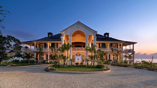 A large, two-story building with a central entrance flanked by columns, surrounded by a landscaped garden and a gravel driveway. - Expansive 6BR Estate Panoramic Sea Views Pool Resort Access (Montego Bay)