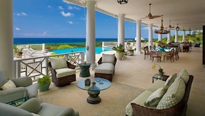 A covered outdoor area with wicker furniture, a round glass table, and a view of the ocean. - Expansive 6BR Estate Panoramic Sea Views Pool Resort Access (Montego Bay)
