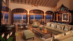 A spacious living room with a large sofa, a coffee table, and a television. There are three arched windows offering a view of the ocean. - Expansive 6BR Estate Panoramic Sea Views Pool Resort Access (Montego Bay)