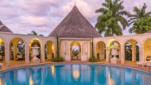 Una piscina con zona techada, paseos con arcos y palmeras al fondo.