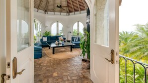 Un umbral abierto que da a un patio cubierto con piso de piedra, ventilador de techo y un espacio de descanso con sofás azules y una mesa de centro.