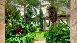Un sendero de jardín que conduce a un pabellón con techo de tejas, rodeado de exuberante vegetación tropical y palmeras.