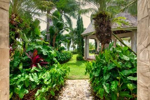A garden pathway leading to a gazebo with a tiled roof, surrounded by lush tropical plants and palm trees.