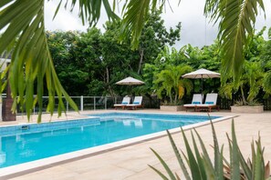 Una piscina con dos sillas de descanso y sombrillas, rodeada de exuberante vegetación.
