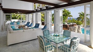 A spacious living area with a glass table, white sofa, and blue chairs, overlooking a pool and palm trees. - Nature-inspired 4BR Modern Villa Pool Resort Access (Montego Bay)