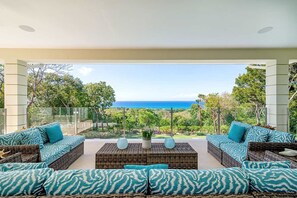 Ein moderner Außenbereich mit Korbmöbeln und gemusterten Kissen, mit Blick auf eine üppige Landschaft und den Ozean.