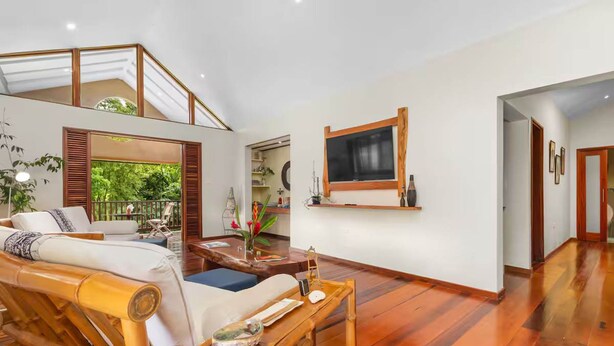 A spacious living area with a high ceiling, a large window overlooking greenery, a seating area with a sofa and a coffee table, and a mounted television. - Hidden 4BR Gem Pool Nature Escape in the Hills of Port Antonio (Port Antonio)