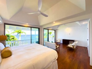 Ein geräumiges Schlafzimmer mit einem großen Bett, einem Deckenventilator und Meerblick durch große Fenster.