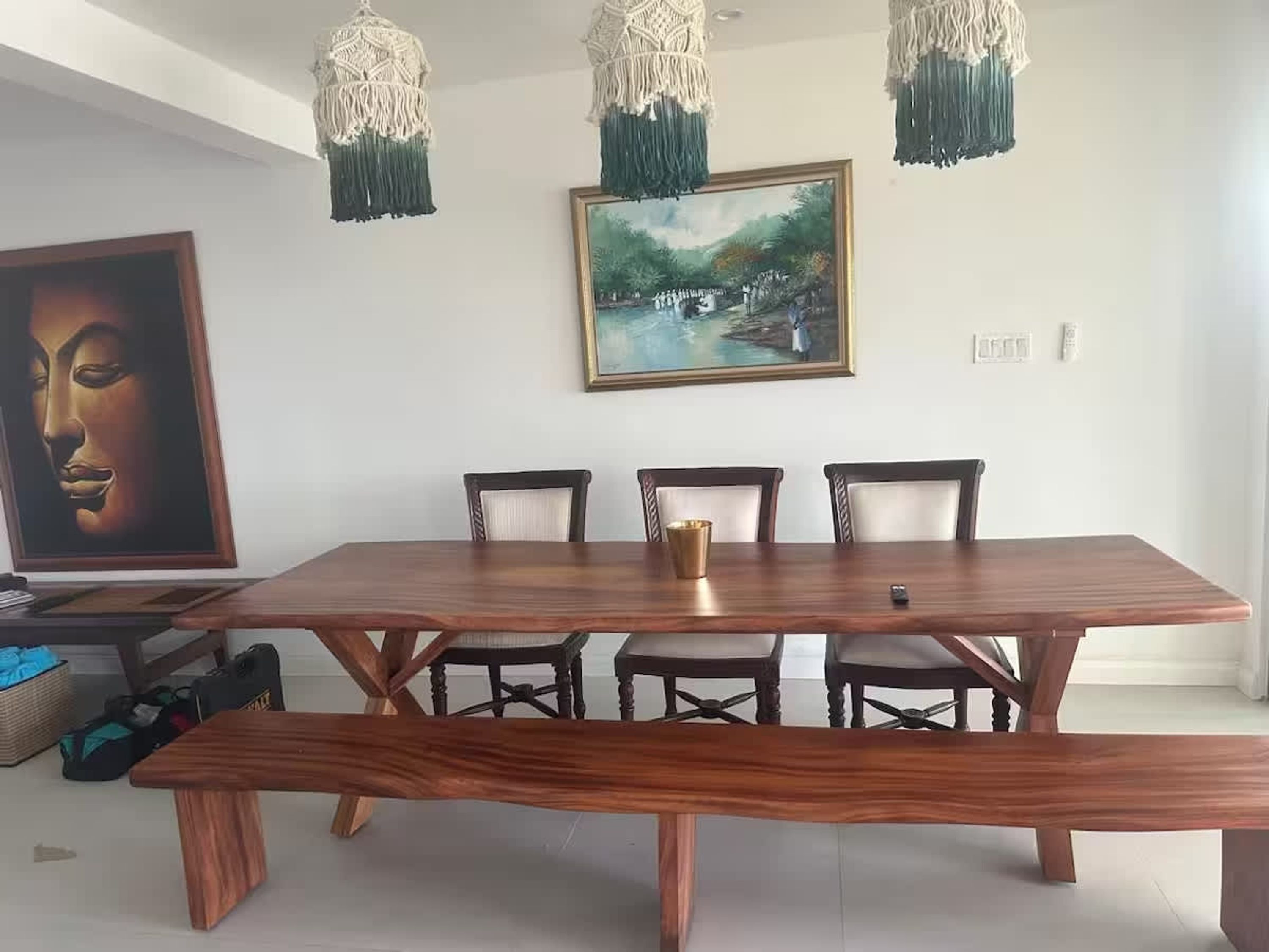 A dining area with a wooden table, four chairs, a bench, macrame hanging lights, and two framed artworks on the wall.