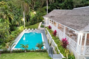Una piscina con cubierta blanca, rodeada de exuberante vegetación y un edificio blanco con tejado de azulejos.