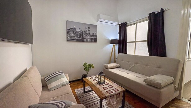 En stue med sofa, lænestol og sofabord. Der er et bybillede på væggen, en aircondition og et vindue med gardiner.