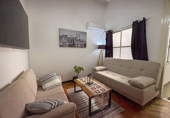 A living room with a sofa, armchair, and a coffee table. There is a cityscape painting on the wall, an air conditioner, and a window with curtains. - Classic, spacious for up to 5 people near Plaza de Mayo and Casa Rosada (Buenos Aires)
