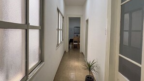 En smal gang med store vinduer, en potteplante og en dør der fører til et rum med en stol og et bord.