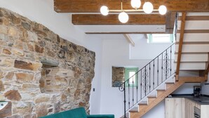 Uma sala de estar ampla com parede de pedra, sofá verde-água, vigas de madeira, escada e área de cozinha.