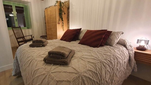 Un dormitorio con una cama grande, una silla de madera, una mesita y una ventana con persianas verdes.