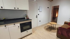 Cocina compacta con gabinetes blancos, encimera negra y horno empotrado. En la sala de estar, hay una pequeña mesa redonda con dos taburetes y un sofá marrón con cojines rojos.