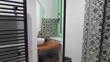 Un baño con ducha, lavamanos e inodoro. Hay un estante de madera con un cuenco y una toalla. Las paredes son blancas y hay una cortina estampada.