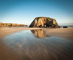 A beach with a large rock formation and calm waters reflecting the sky.