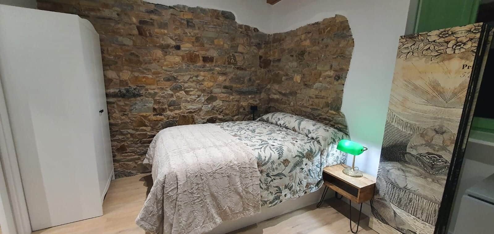 Quarto com parede de pedra, cama com roupa de cama florida, mesa de cabeceira com abajur e um quadro grande na parede.