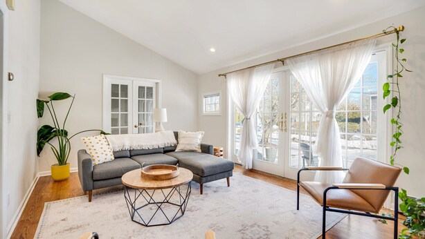 Ein modernes Wohnzimmer mit einem grauen Sofa, einem Holztisch und einem braunen Ledersessel. Es gibt ein großes Fenster mit weißen Vorhängen, einen Blumentopf und eine Lampe auf einem Beistelltisch.