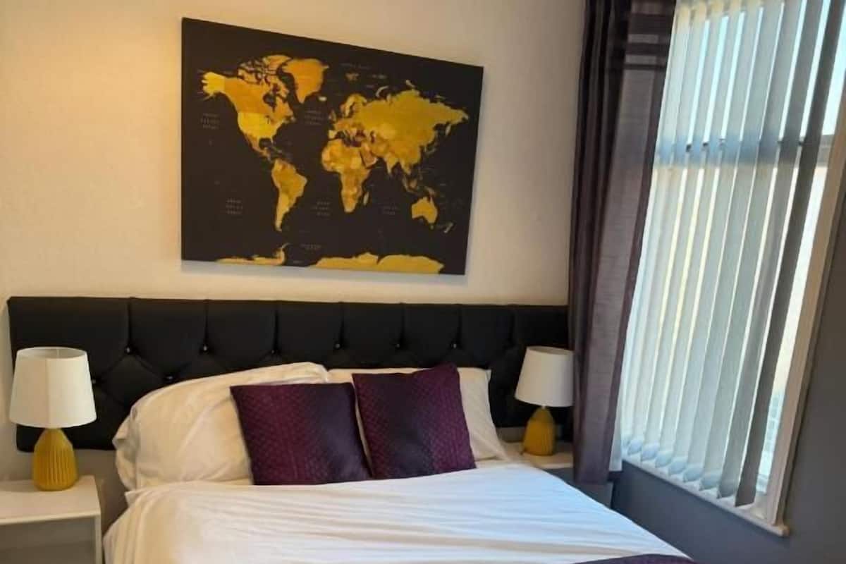A hotel room with a large bed, two bedside tables with lamps, a world map on the wall, and a window with blinds.