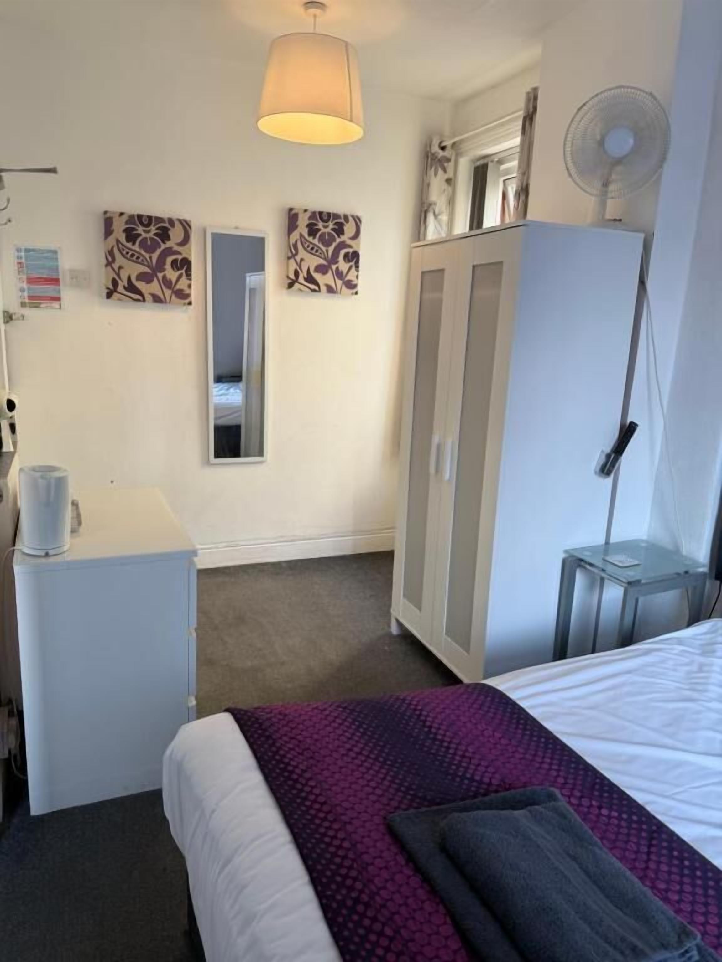 Una habitación de hotel con cama, escritorio con lámpara, armario y ventilador de pie.