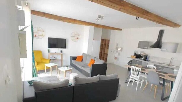Ein modernes Wohnzimmer mit einem grauen Sofa, einem gelben Sessel und einem Fernseher. Es gibt eine Küchenzeile mit einem Herd und einem Esstisch.