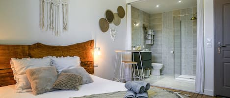 A bedroom with a wooden headboard, a bed with rolled towels, a bedside table, a wall-mounted dreamcatcher, and a bathroom with a shower and toilet visible through an open door.