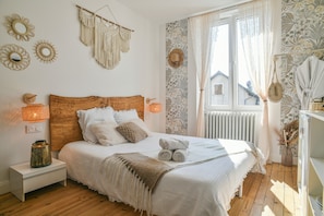 A bedroom with a wooden headboard, a bed with white bedding, a nightstand with a lamp, a radiator, and a window with curtains. - Boho Room with Garden and Private Jacuzzi (Villages du Lac de Paladru)