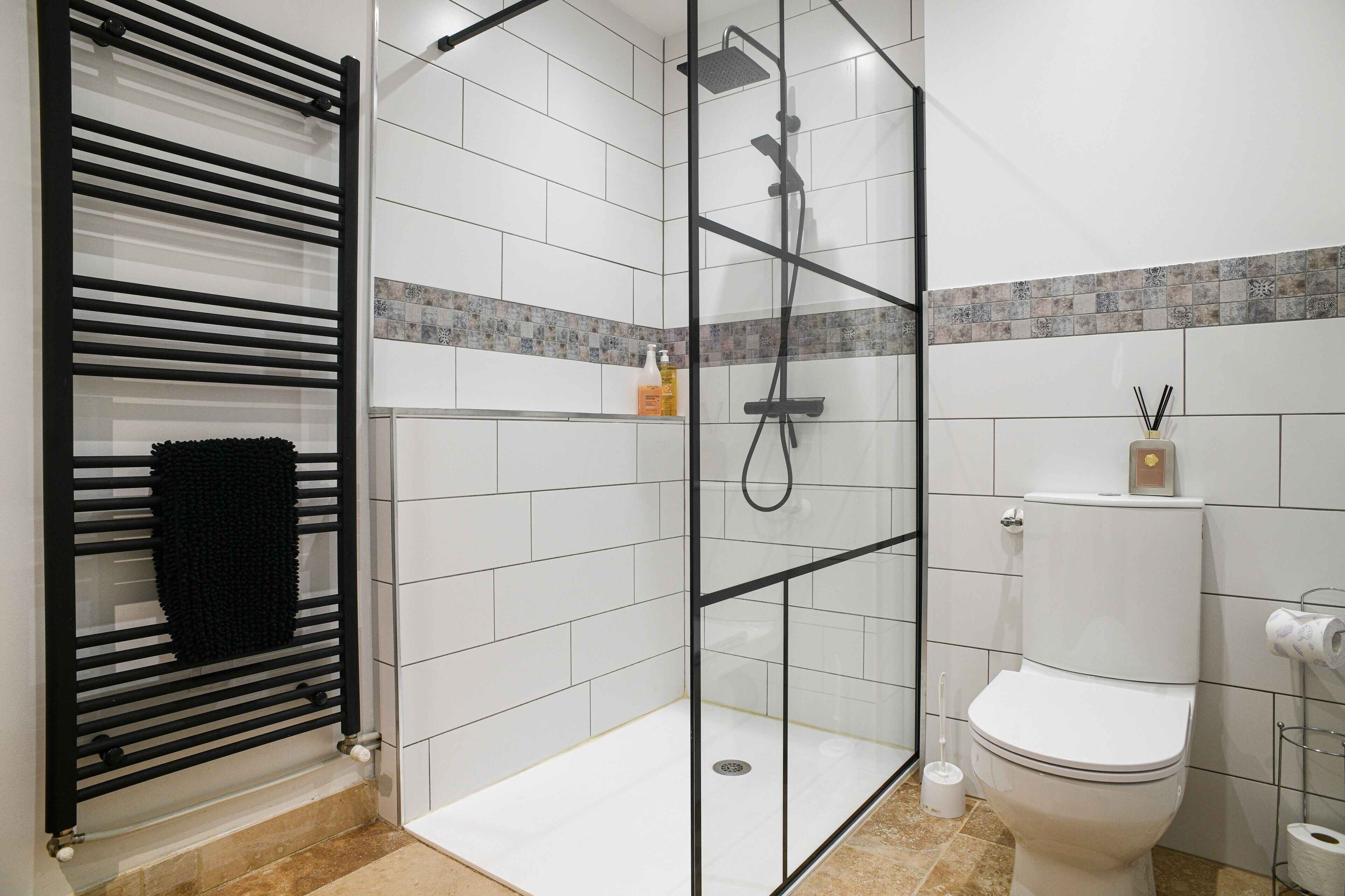 Un baño moderno con ducha envuelta en vidrio, toallero negro, inodoro y salpicadero de azulejos.
