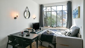 Een kamer met een bed, een bureau met een computer, een stoel, een tafel met een drankje en een raam met gordijnen.